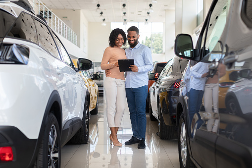 Young Black Couple Choosing Car In Modern Dealership Center, Holding Clipboard And Checking Vehicle Characteristics In Catalog, Happy African American Spouses Buying Automobile In Showroom