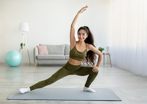 Sporty Indian woman stretching her body during home workout, copy space. Full length of fit young Eastern lady training indoors, leading active lifestyle during covid lockdown