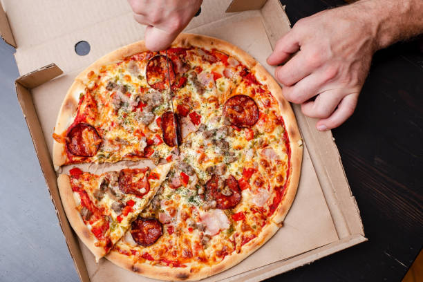 istock Man hand is cutting an appetizing pizza into slices, in a box, on a black table. Top view. 1354209600