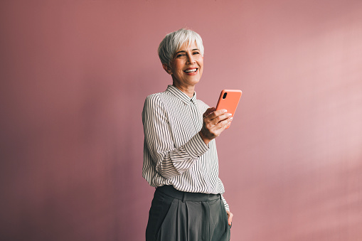 Smiling businesswoman standing over a pink background and typing text message on her smartphone and looking at camera.
