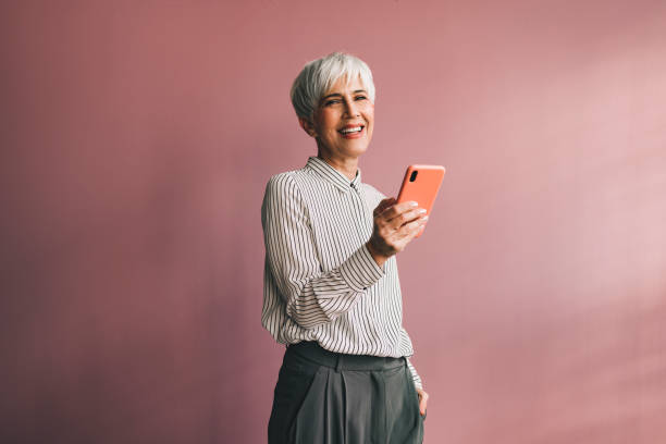 Portrait of a Senior Business Woman Using Mobile Phone Smiling businesswoman standing over a pink background and typing text message on her smartphone and looking at camera. brand name smart phone photos stock pictures, royalty-free photos & images