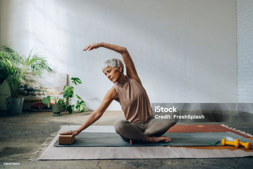 Senior Woman Doing Home Workout Training Serious woman in sportswear sitting on a exercise mat and stretching arms. Yoga Stock Photo