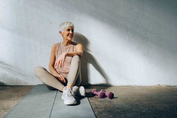 Smiling Senior Woman Sitting on a Exercise Mat Happy woman in sportswear looking away while resting after home workout training. studio yoga adult action stock pictures, royalty-free photos & images