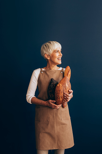 Smiling female baker holding baked breads in hands while standing over a blue background.