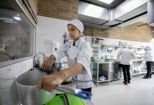 Back view of a male chef draining noodles by the kitchen sink. He is wearing black gloves and uniform.