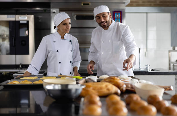 бейкер учит своего ученика, как делать хлеб в пекарне - hat women chef occupation стоковые фото и изображения