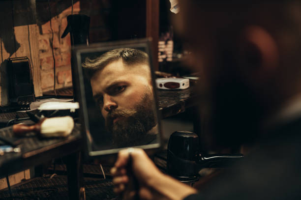 homme se regardant dans le miroir alors qu’il est assis sur une chaise dans un salon de coiffure - one man only human age mustache beard photos et images de collection