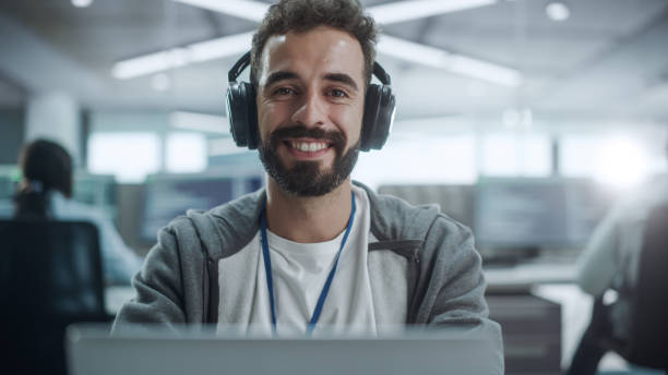 istock Office: Portrait of Happy IT Programmer Wearing Headphones Working on Desktop Computer, Looking at Camera and Smiling. Male Software Engineer Developing App, Video Game. Listening to Podcast, Music 1354214671