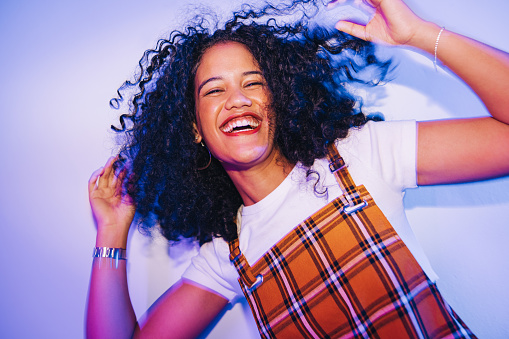 Young woman dancing and whipping her hair happily. Cheerful young woman laughing joyfully while standing alone in bright neon light. Carefree young woman having having fun at a house party.