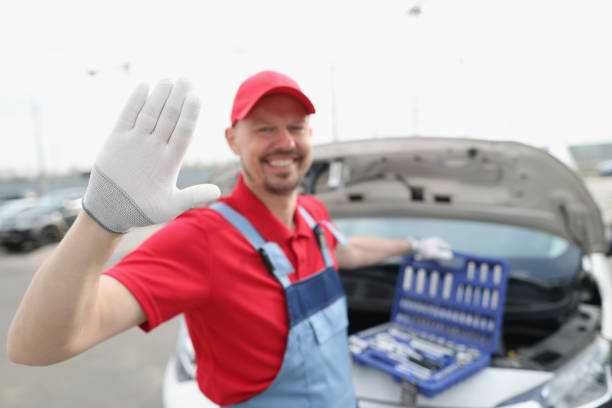 Man in a safety vest works on a car engine with scenic mountains in the background