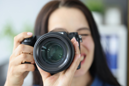 Close-up of smiling brunette female photographer pointing super wide lens at you. Girl professional photographer with camera. Art, creative, studio concept