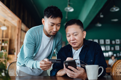 Joyful Asian senior father and son shopping online with mobile app on smartphone together and making payment with credit card online at home