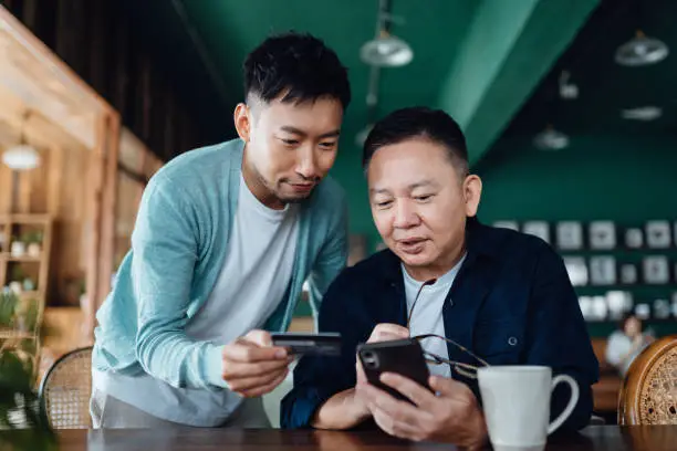 Photo of Joyful Asian senior father and son shopping online with mobile app on smartphone together and making payment with credit card online at home