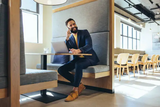 Cheerful young entrepreneur speaking on a phone call. Happy young businessman smiling while communicating with his business partners. Successful entrepreneur working in a modern workspace.