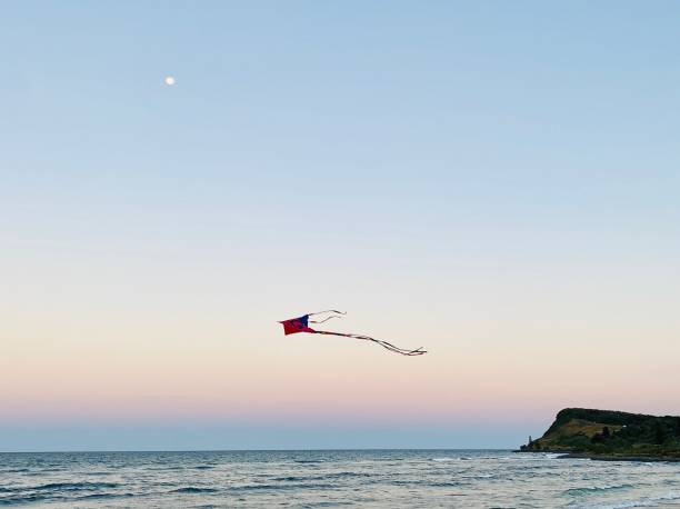 Kite Below Full Moon at Beach Sunset Horizontal seascape of pink children’s kite sailing high in the sky above ocean waves with background coastline headland and full moon at sunset Lennox Head beach NSW Australia kite sailing stock pictures, royalty-free photos & images