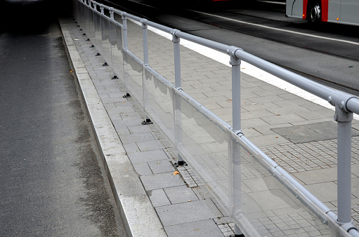 the bus platform is protected on one side by transparent plexiglass attached to a gray metal tube railing. People are not sprayed with dirt from passing cars
