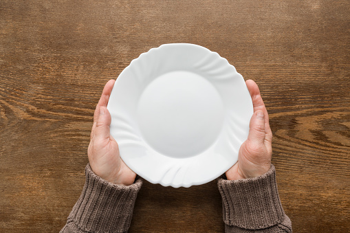 Mature woman hands holding white empty plate on dark brown wooden table background. Closeup. Point of view shot. Meal waiting concept. Top down view.