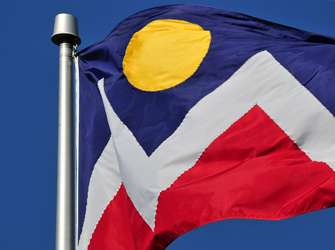 Denver, Colorado, USA: official flag of the City and County of Denver in Colorado. It was designed by a North High School student, Margaret Overbeck, and adopted in 1926. A zigzag white stripe horizontally separates a red field below from a blue field above, in which is centered a yellow circle, together forming a stylized depiction of the Sun in a blue sky above snow-capped mountains. Waving in the wind.