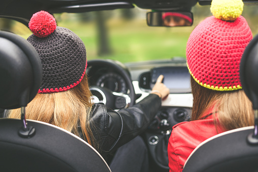 Trendy girls sitting in the car. Teen in front of the steering wheel with a friend beside. Young fashion women enjoying free time in a convertible car. Youth, freedom, travel and happiness concept.