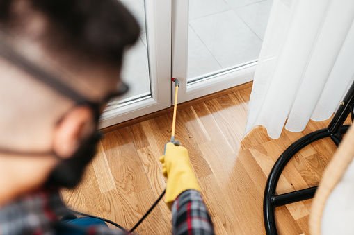 Professional exterminator in protective workwear spraying pesticide in apartment.