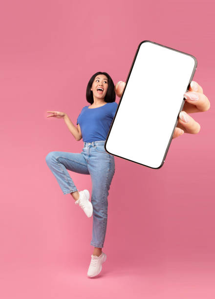 istock Excited young Asian lady jumping, shouting OMG, presenting smartphone with empty screen over pink background, mockup 1354242720