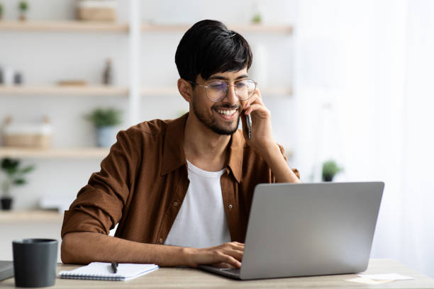 cheerful indian guy businessman with laptop having phone conversation - minority professional occupation business ethnic imagens e fotografias de stock