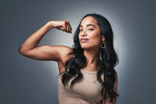 studio shot of a beautiful young woman flexing while standing against a grey background - flexing muscles fotos imagens e fotografias de stock