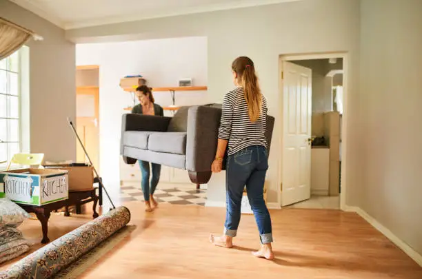 Two young female friends carrying a sofa out of a house together while moving to a new home