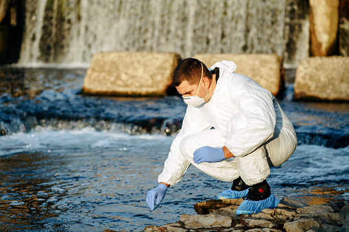 Environmental inspector in white safety suit examining polluted river water at industrial site