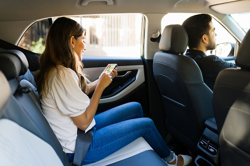 Side view of an attractive latin woman sitting on the car back seat and using a ride share app on her smartphone