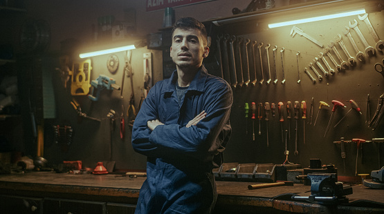 Serious handsome blacksmith man with workwear fold one's arms, looking at camera on awesome workshop background