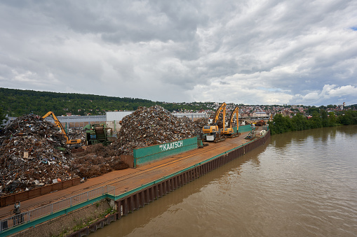 Plochingen, Germany - July 16, 2021: Company Kaatsch scrap metal trade and recycling. Heap with old iron. Grey sky and brown river water. Taken from a bridge.