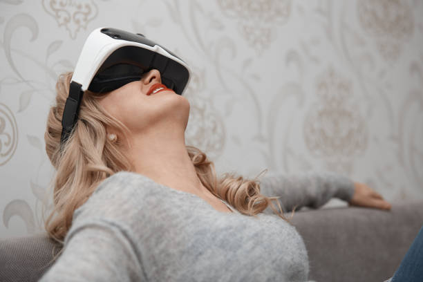 istock Blonde smiling woman wearing Virtual reality headset while lying on the sofa 1354260813