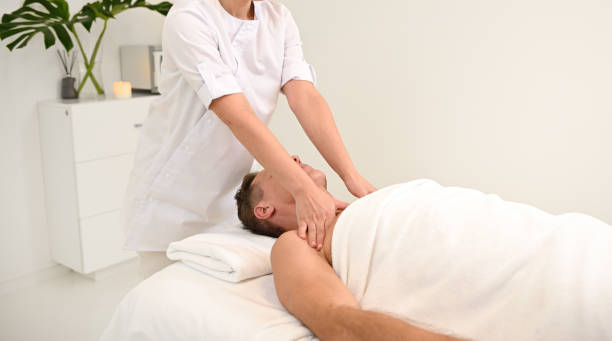Unrecognizable masseuse giving shoulder massage to a middle aged man relaxing on massage table in luxury white spa beauty clinic. Therapeutic body massage treatment, body care concept with copy space masajista irreconocible dando masaje de hombros a un anciano que se relaja en la mesa de masajes en la lujosa clínica de belleza de spa blanco. tratamiento terapéutico de masaje corporal, concepto de cuidado corporal con espacio de copia