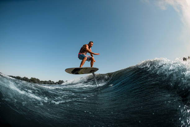 uomo atletico in equilibrio sull'onda con lavagna aliscafo su sfondo di cielo blu - wakeboarding surfing men vacations foto e immagini stock