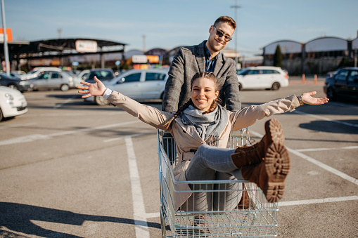 Full body crazy man and cheerful woman looking at each other while riding shopping cart on asphalt parking lot