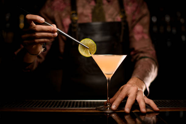 martini glass with cocktail on bar and hand of bartender decorates the glass with slice of lemon - transparent holding glass focus on foreground imagens e fotografias de stock