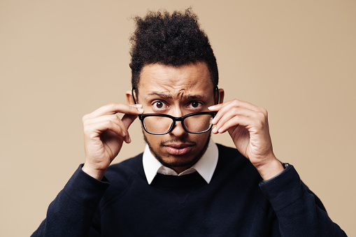 Young worried Afro man looking over glasses on beige background. Face expression, emotions concept