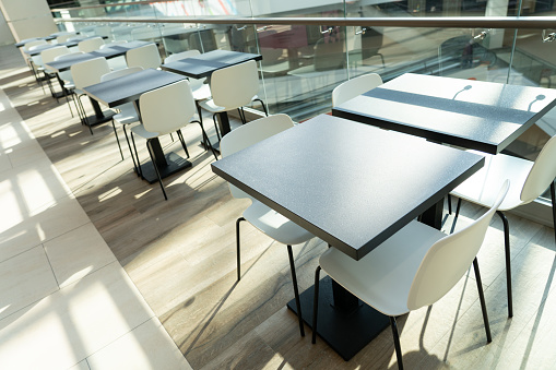 A row of tables with white chairs for visitors to the food court of a modern shopping center. People admire the beautiful view during the meal.