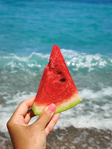 Close up. Female hand holding a slice of juicy red watermelon against the background of the sea / ocean. Vertical oriented.