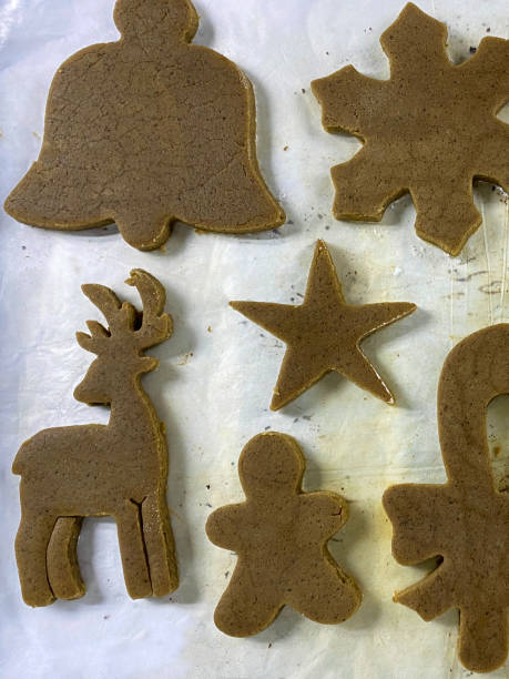 Stock photo showing close-up, elevated view of greaseproof parchment paper with cookie dough cut out into uncooked, homemade gingerbread Christmas themed biscuits ready to bake in the oven. Home baking concept photo. imagen en primer plano de galletas navideñas sin cocinar, caseras, de pan de jengibre en papel de pergamino a prueba de grasa, campana, copo de nieve, estrella, reno, hombre de pan de jengibre y caña de caramelo cortadas formas, horneado casero, vista el