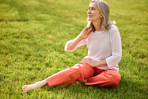Time for yourself. Adult gray-haired good looking woman in casual clothes barefoot sitting on grass in park looking thoughtfully aside touching her hair