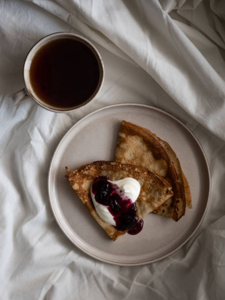 istock Breakfast in bed, a cup of black tea and thin pancakes with currant jam and sour cream on a beige ceramic plate on a soft white bed 1354354084