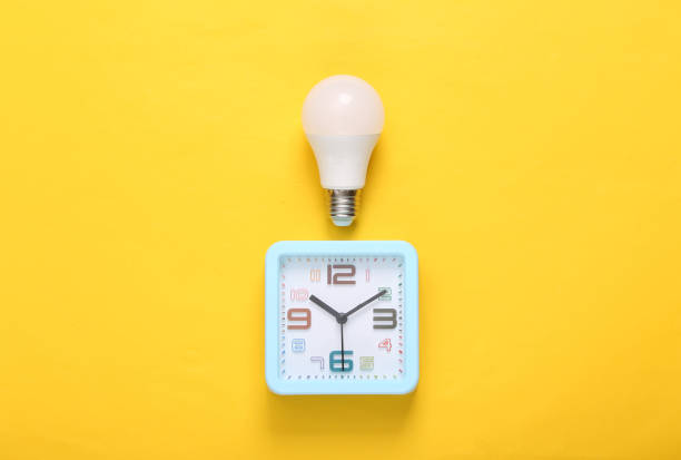 istock Alarm clock and light bulb on yellow background. Top view 1354361262