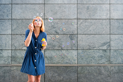 Carefree young female in blue dress blowing rainbow soap bubbles while having fun in summer day against gray wall background
