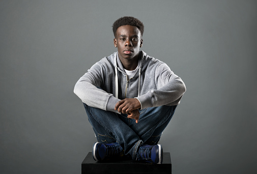 Young Black man in grey hoodie and jeans posing cross-legged in a studio looking at the camera with a serious deadpan expression