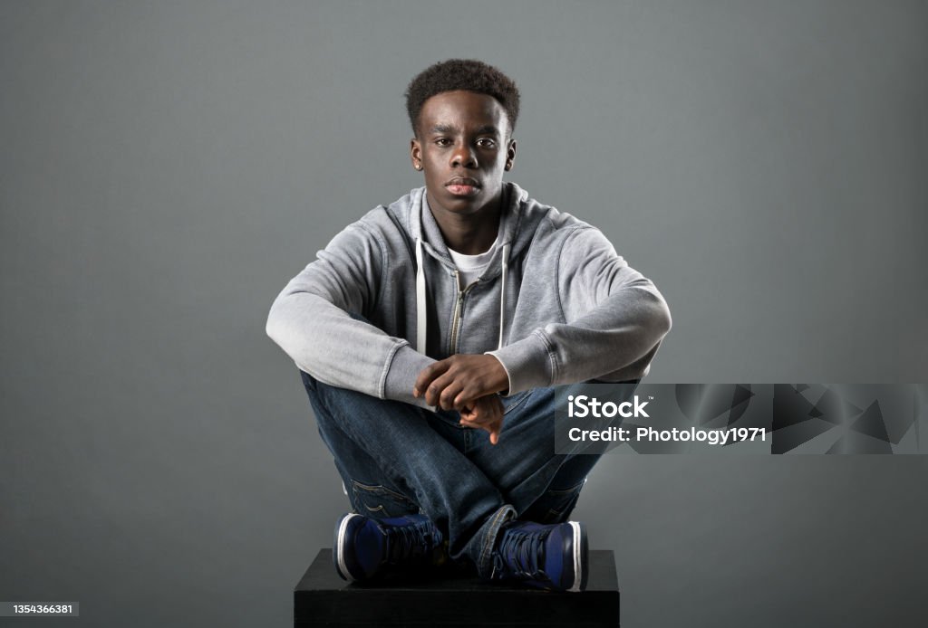Young Black man posing cross-legged in a studio Young Black man in grey hoodie and jeans posing cross-legged in a studio looking at the camera with a serious deadpan expression Teenager Stock Photo