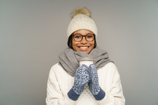 Young black woman in warm hat and mittens hold paper coffee cup. Female wearing winter woolen scarf and knitted sweater drink hot chocolate or tea from disposable takeaway mug warming up in wintertime