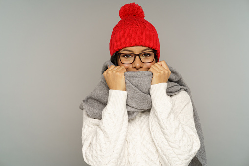 Funny woman in glasses, sweater, knitted hat and scarf covering face. Joyful african american female wear warm clothes for cold weather and frost. Isolated studio portrait of cute cheerful black girl.