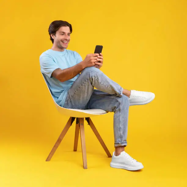 People And Technology Concept. Portrait of smiling young man using smartphone sitting on chair isolated on orange studio background. Excited casual guy chatting online, browsing social media